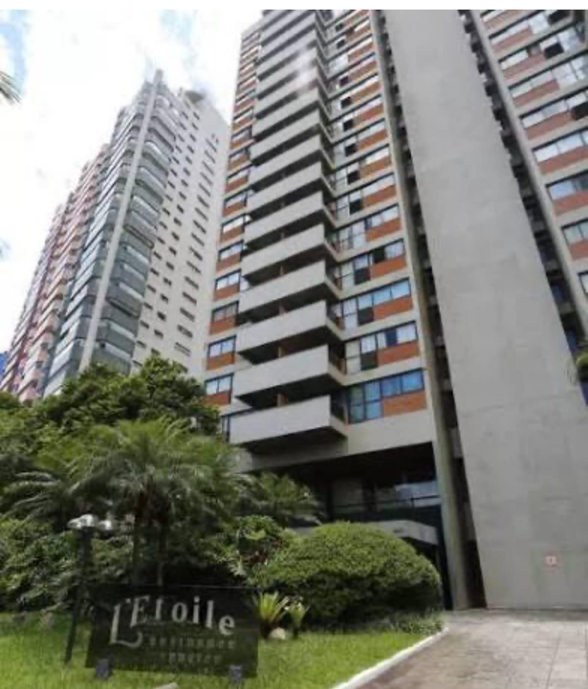 A tall apartment building is showcased, featuring a modern facade with a mix of glass and concrete elements. Lush greenery surrounds the entrance, providing a refreshing contrast to the building's architecture. The name 'Etoile' is elegantly displayed at the base, inviting guests to the residence.