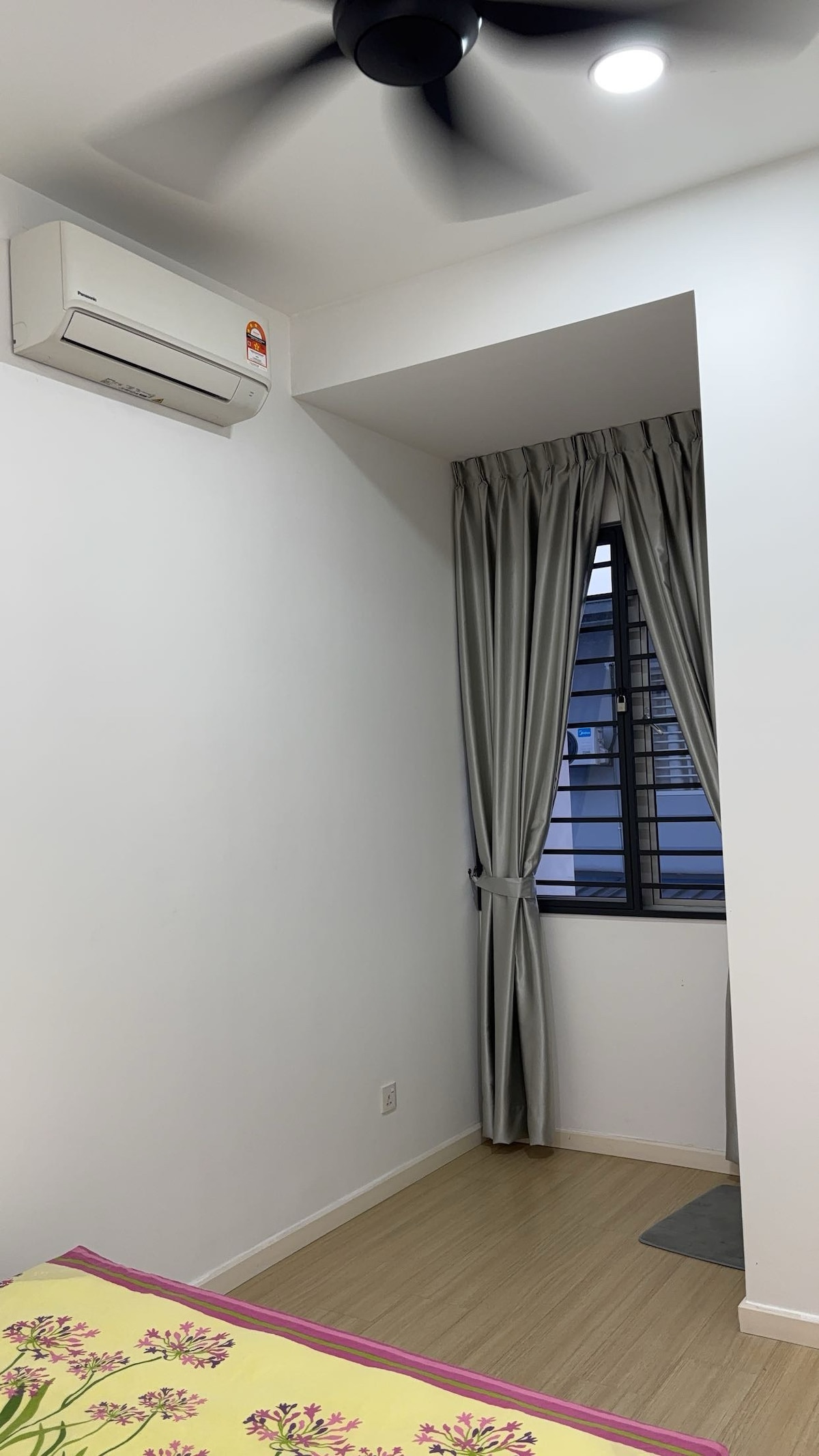 A bright room features a sleek air conditioning unit mounted on the wall. Natural light filters through a window adorned with gray curtains. Smooth wooden flooring complements the minimalist design, creating an atmosphere of simplicity and comfort.