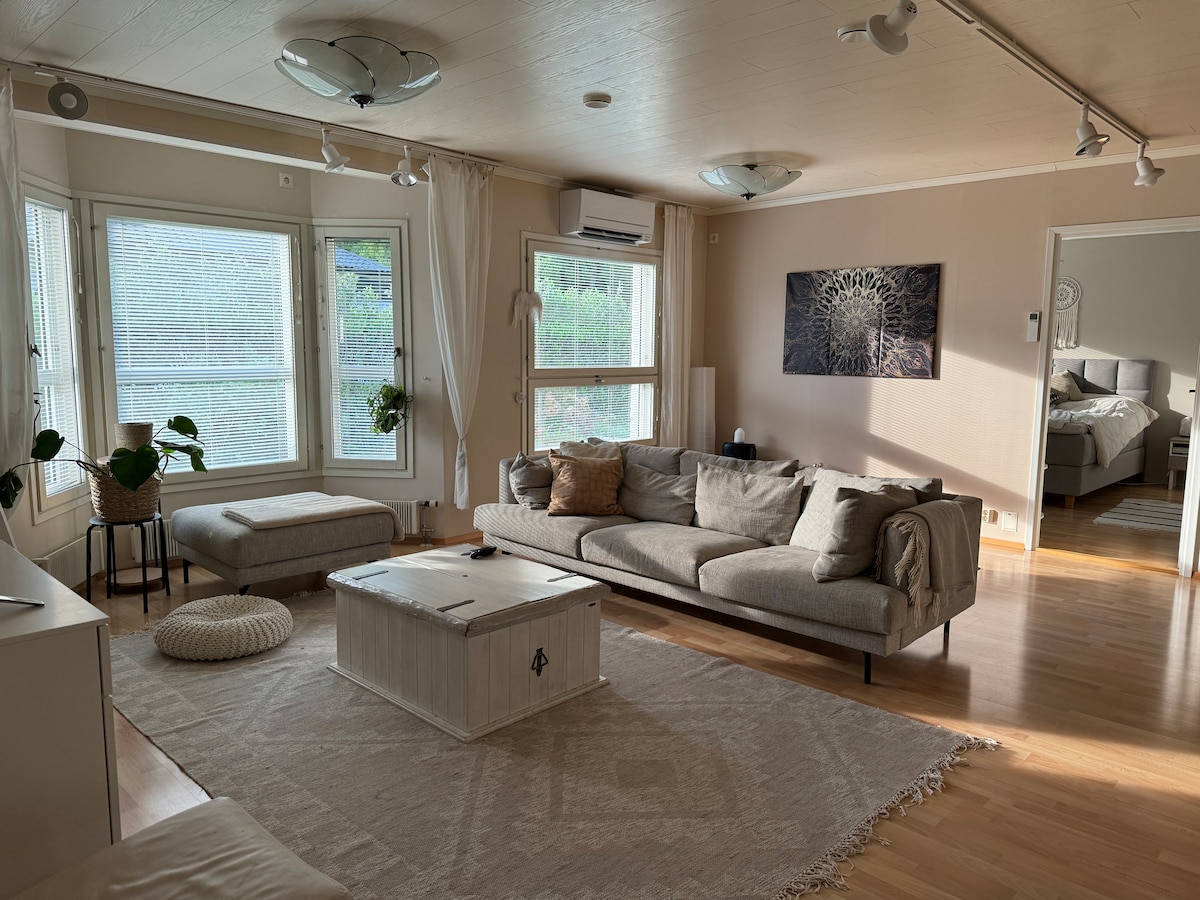 A spacious living area features a light-colored sofa arranged around a central coffee table. Large windows allow natural light to fill the room, complemented by modern ceiling lights. A cozy seating option is positioned in the corner, and a view of the adjacent bedroom is visible.