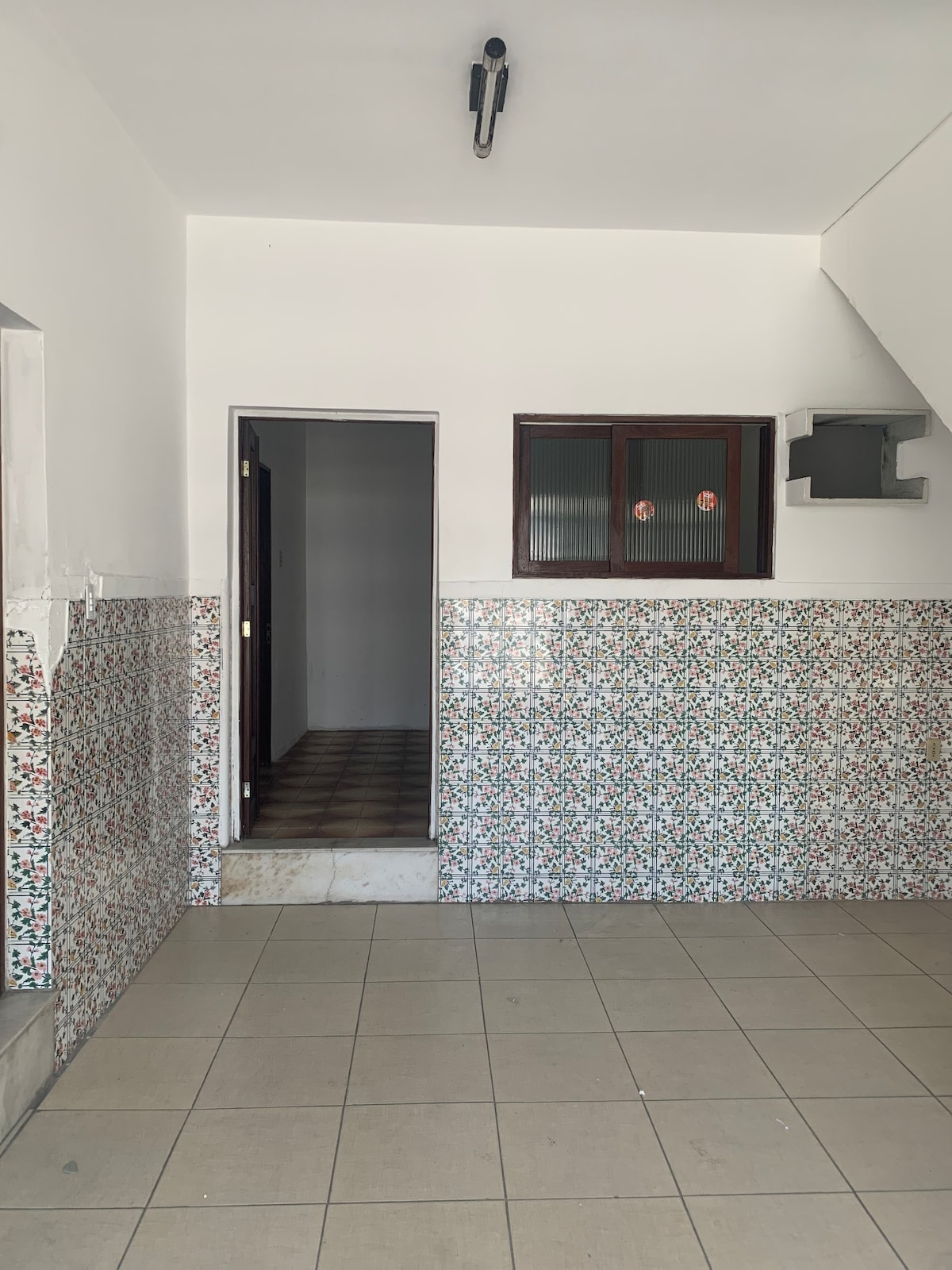 A well-lit interior space features tiled flooring and colorful patterned wall tiles. A doorway leads to a darker area beyond, while a window allows natural light to enter. The simplicity of the design enhances the versatility of the room for various uses.