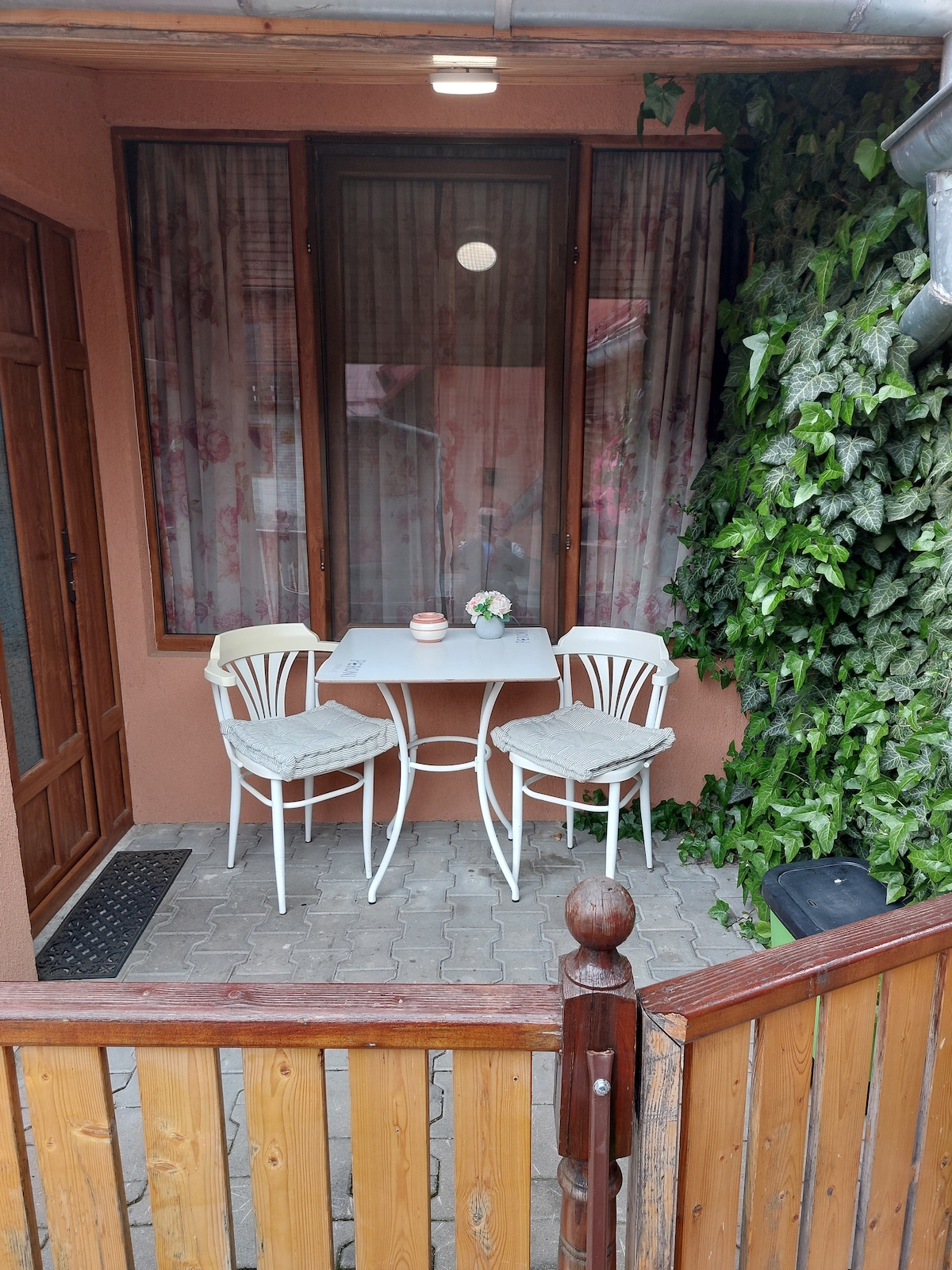 A small outdoor terrace is featured, with a rectangular table and two chairs. The area is framed by greenery, and light curtains filter the view. A door is visible on the left, leading into the interior space.