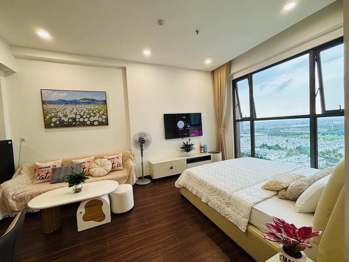 A bright and airy living space features a spacious bed and a comfortable sofa set. A large window offers views of the surrounding area. A flat-screen television is mounted on the wall opposite the seating area, while a small coffee table adds functionality.