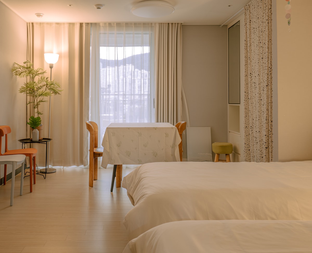 A spacious room features two beds with crisp white linens, complemented by a decorative table set for dining. Natural light floods in through large windows, draped with light curtains, while a floor lamp and potted plant add subtle warmth to the atmosphere.