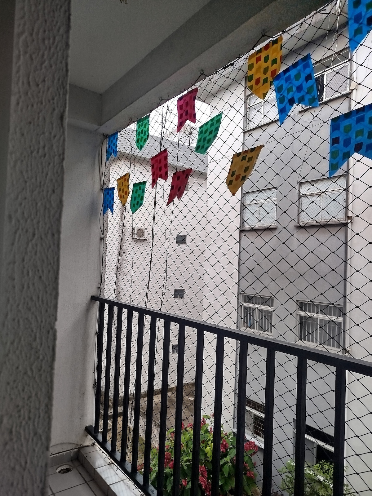 A balcony space features a decorative net with colorful flags in red, blue, yellow, and green. The view includes gray buildings and partially visible greenery, providing a lively atmosphere. A railing adds safety and structure to the area, enhancing the outdoor experience.