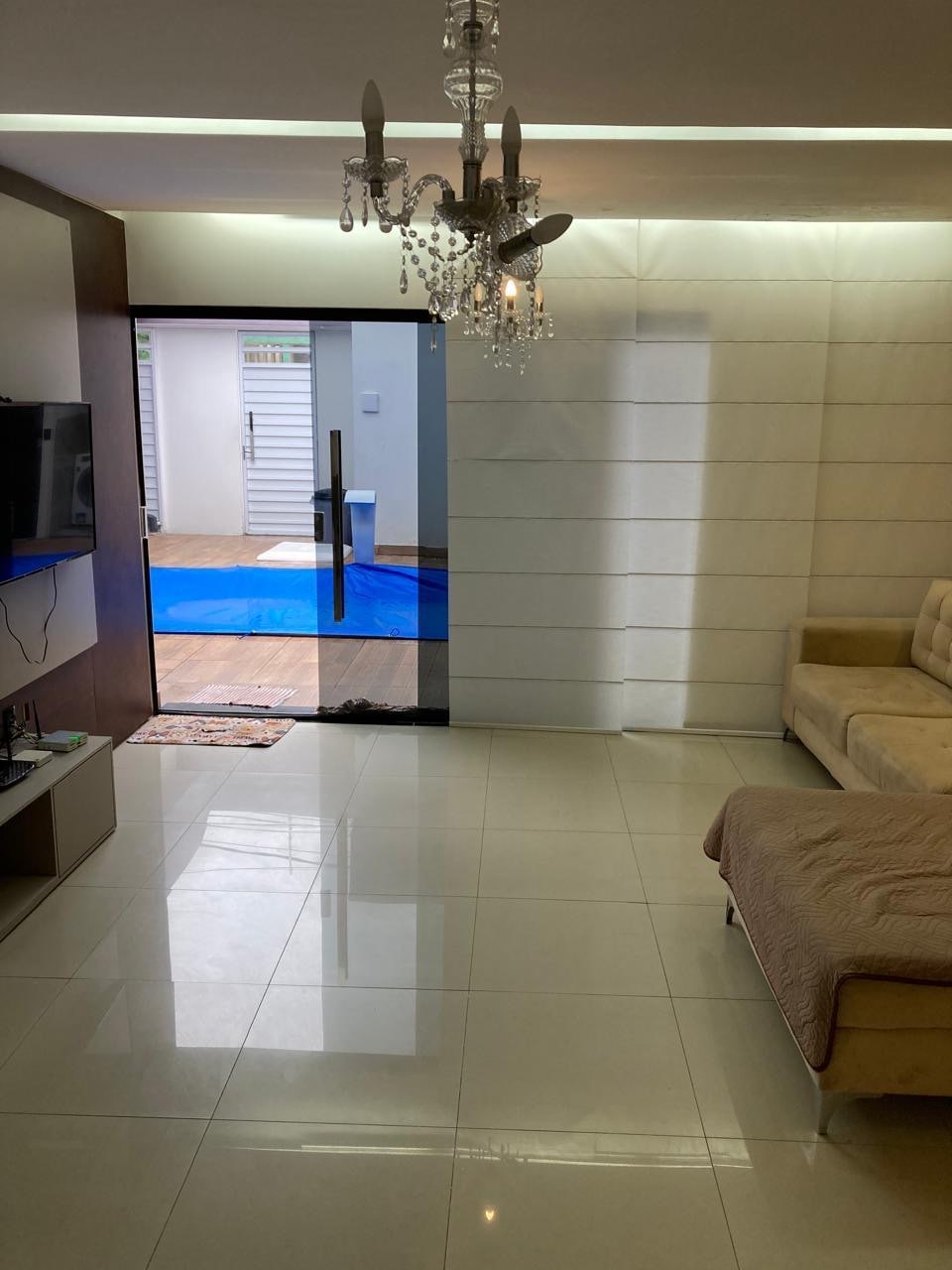 A spacious living area features glossy white tile flooring and a contemporary chandelier. A light-colored sofa is positioned against the wall. Sliding glass doors lead to an outdoor space, allowing natural light to fill the room.