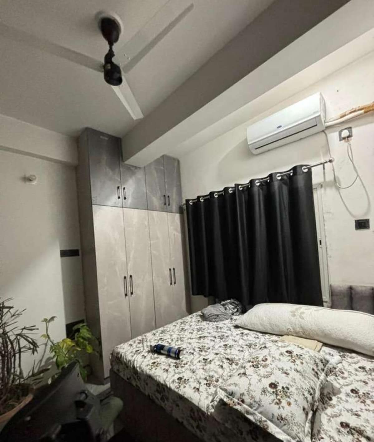 A cozy bedroom features a spacious bed with floral bedding, complemented by a cushioned headboard. Dark curtains frame a window, while a ceiling fan provides ventilation. A wardrobe stands against the wall, and a small indoor plant adds a touch of greenery to the space.