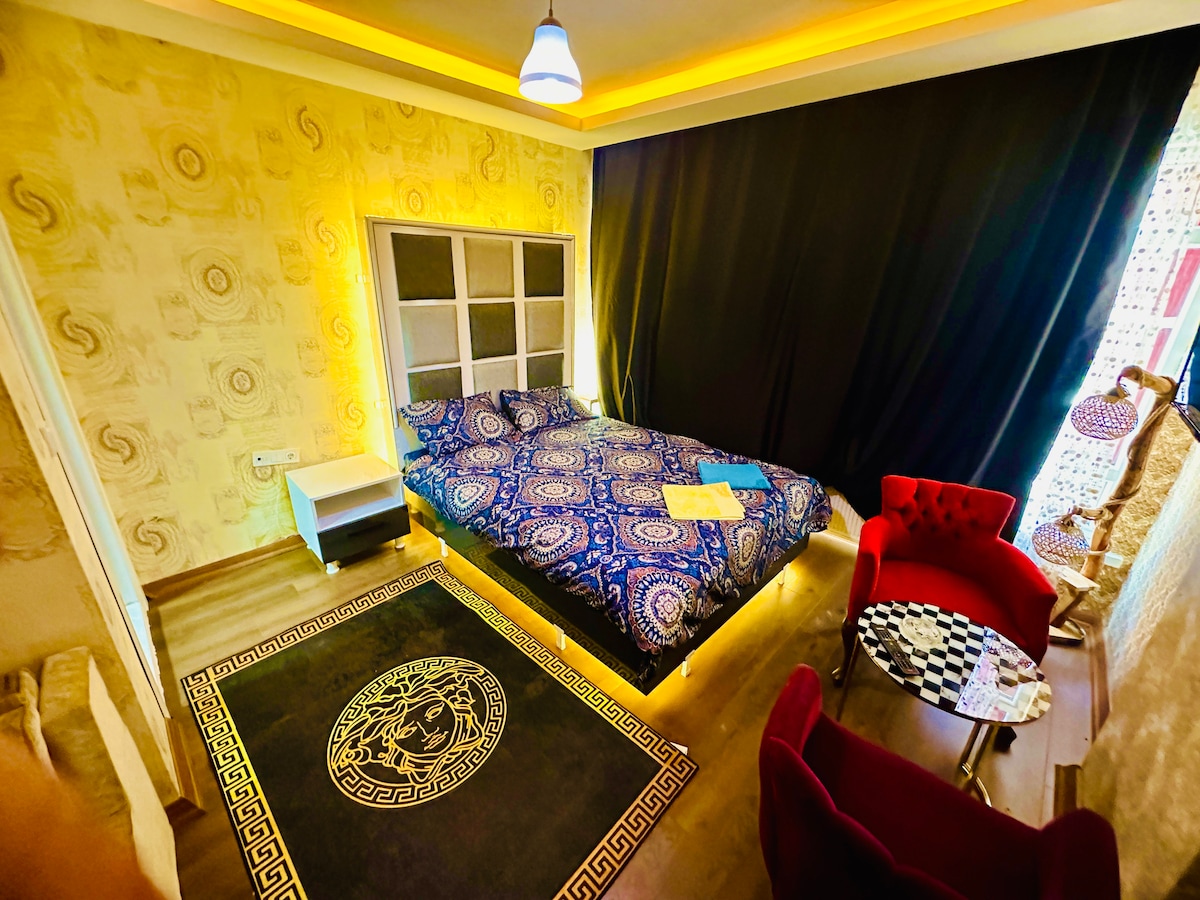 A spacious sleeping area is presented with a large bed featuring a vibrant patterned bedspread. A dark curtain partially covers a window, and a cozy seating area showcases a red chair and a small table. A decorative rug enhances the room's design with a geometric pattern.