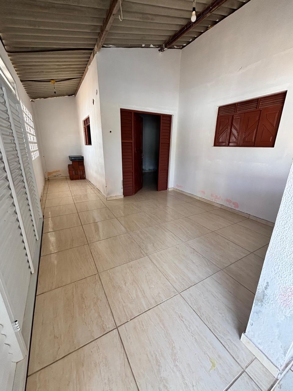 A spacious corridor is shown with tiled flooring and white walls. Light enters through two small brown-framed windows, and a wooden cabinet is positioned at the far end. Doorways lead to adjacent rooms, creating an open and airy atmosphere.