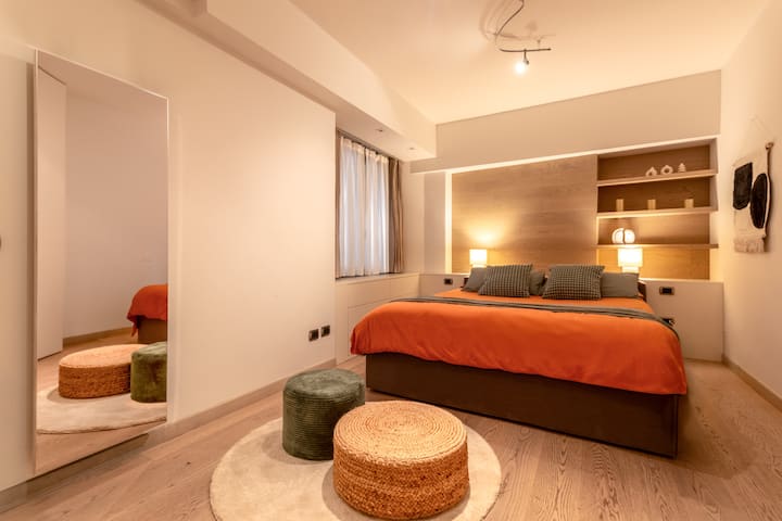 วิวห้องนอนหลักที่มีพรมอยู่ตรงกลางซึ่งมีเก้าอี้สองตัวสร้างพื้นที่พักผ่อนที่สง่างามด้านหน้ากระจกติดผนังแนวตั้งขยายพื้นที่อย่างเห็นได้ชัดและสะท้อนแสง