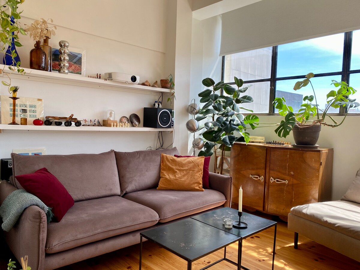 A cozy living area features a comfortable brown sofa adorned with decorative cushions. A small black coffee table sits in front, while green plants add a refreshing touch. Natural light fills the space through large windows, illuminating decorative shelves and wooden accents.