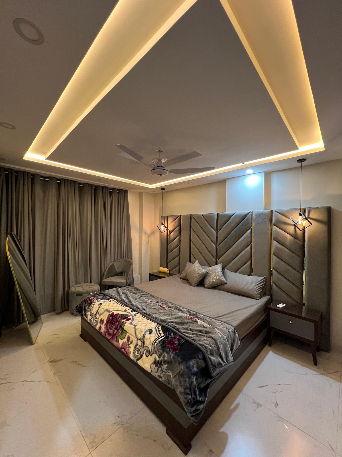 A modern bedroom is presented, featuring a spacious bed with decorative pillows against a textured headboard. Soft lighting accentuates the coffered ceiling, while floor-to-ceiling, draped curtains frame a glass window. A stylish chair and a small bedside table complement the serene environment.
