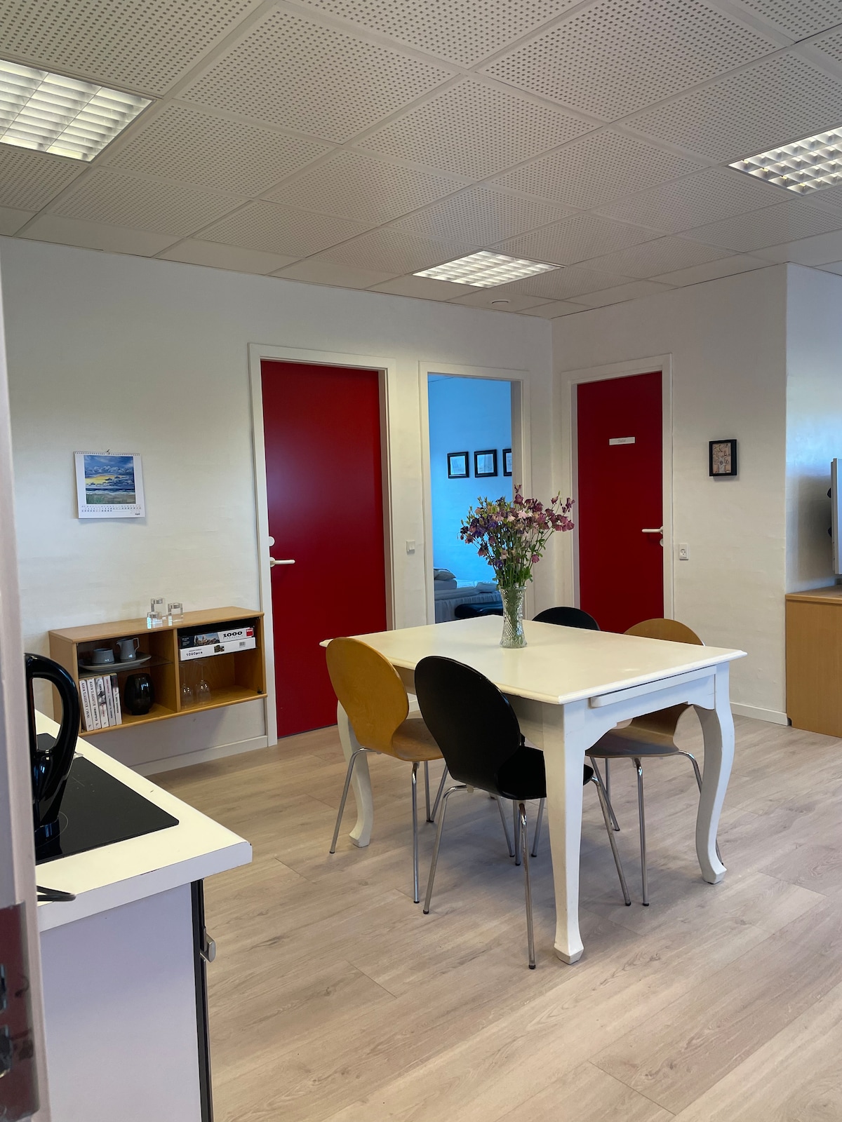 A bright dining area features a white table with four chairs in varied colors. Doors in deep red provide contrast to the light walls. A small shelf displays a few decorative items, and natural light illuminates the space, creating a welcoming atmosphere.