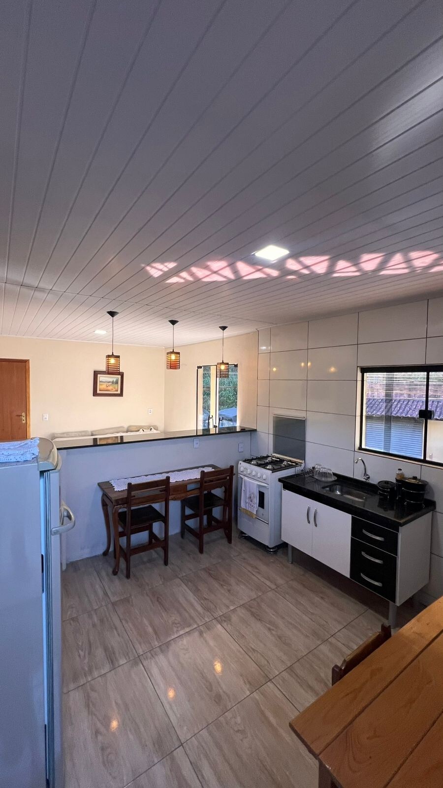 A functional kitchen area is shown with modern cabinetry and appliances. A dining table with four chairs is situated near a window, allowing natural light to fill the space. The ceiling is white with recessed lighting, contributing to a bright atmosphere.