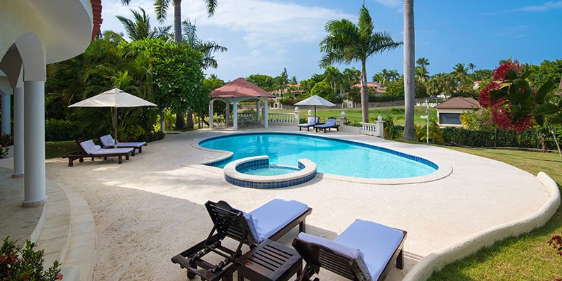 A private swimming pool is surrounded by a spacious terrace with lounge chairs and umbrellas. Lush tropical gardens with palm trees and vibrant foliage frame the pool area, offering a tranquil outdoor setting. A shaded pavilion is visible in the background.