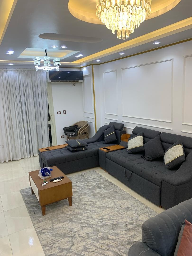 A modern living area is featured, showcasing dark gray sectional seating with decorative cushions. A wooden coffee table rests on a patterned rug, and a chandelier provides ambient lighting. Sheer white curtains frame large windows, enhancing the space's brightness.