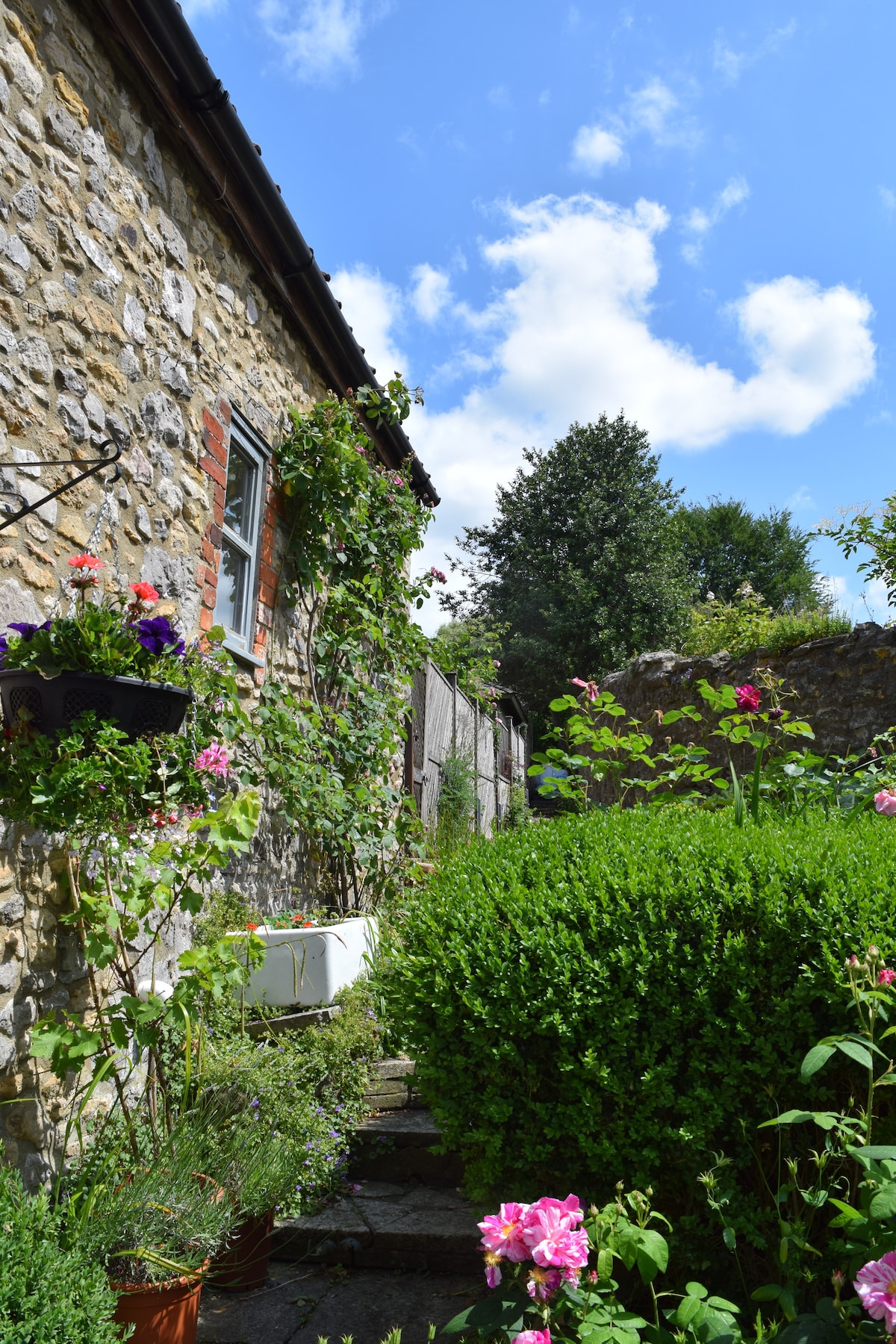 The exterior of the cottage features charming stone walls adorned with climbing plants and colorful flower boxes. A well-maintained path leads alongside lush green hedges, inviting guests towards the entrance. The bright sky adds a sense of openness to the tranquil garden setting.