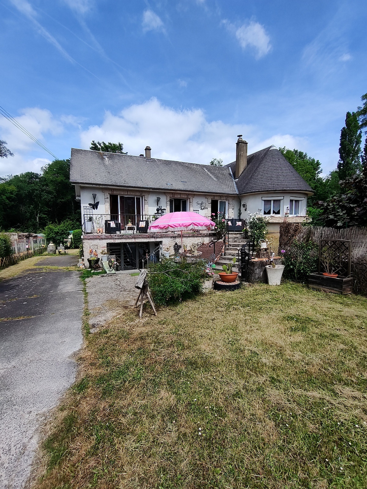 The exterior of the house features a light-colored façade with multiple windows and a gently sloped roof. A pink parasol is positioned over an outdoor seating area, surrounded by decorative plants. An expansive lawn leads to the entrance, with a gravel pathway visible.