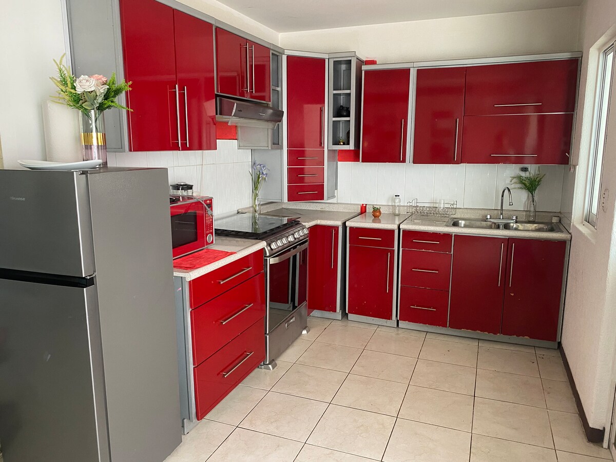 A modern kitchen features bright red cabinetry complemented by stainless steel appliances, including a refrigerator and a gas stove. A countertop with a sink provides ample workspace, while natural light enters through a nearby window, enhancing the functional layout.