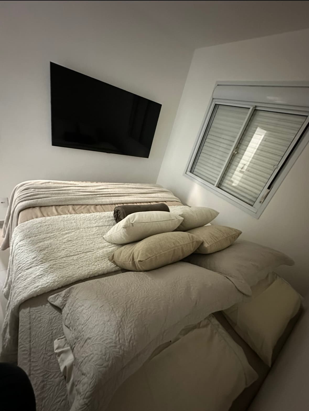 A snug sleeping area is presented with layered bedding in neutral tones. Multiple pillows are arranged on a spacious bed, complemented by a sizable window that allows natural light to enter. A flat-screen television is mounted on the wall opposite the bed.