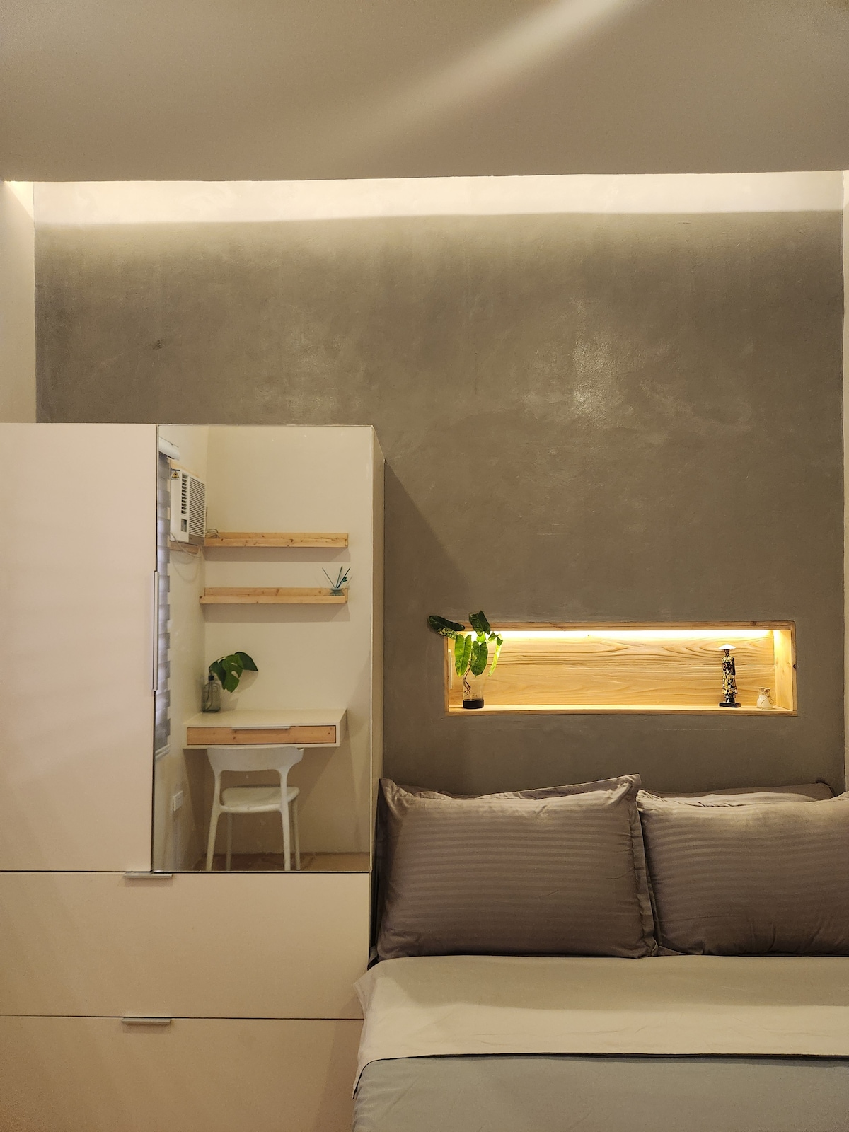 A minimalist bedroom features a neutral grey accent wall and sleek furnishings. A small workspace with a chair and decorative plants is positioned beside a built-in shelf. Soft pillows adorn the spacious bed, complemented by warm lighting from a recessed area showcasing additional greenery.