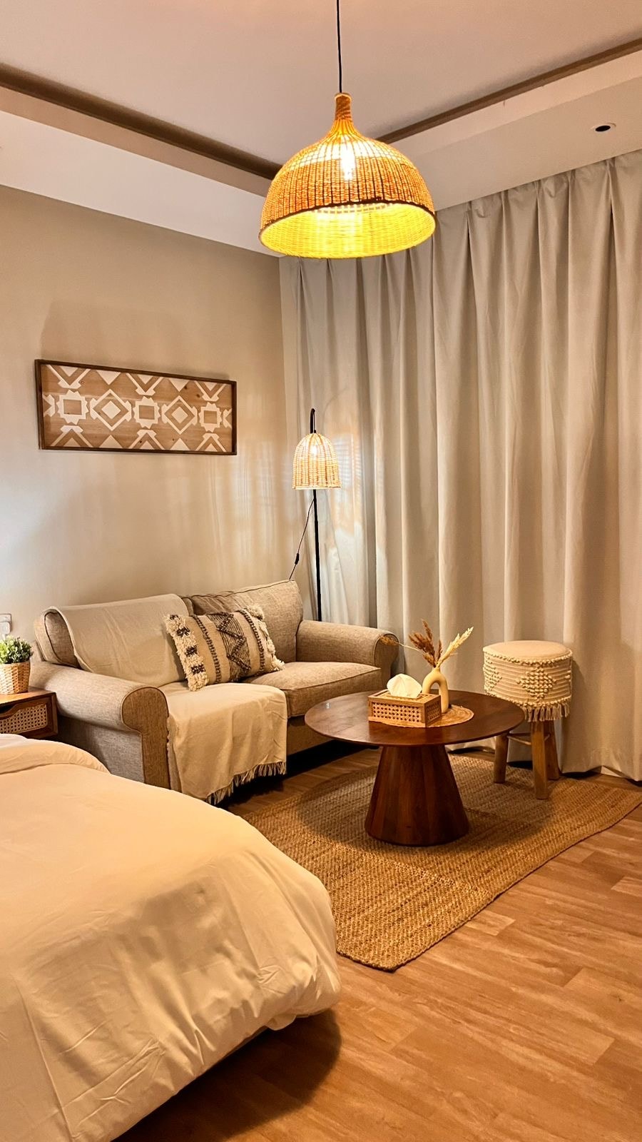 A cozy corner of the studio is highlighted by a comfortable seating area, featuring a plush sofa with decorative cushions. A round wooden table is positioned nearby, accompanied by stylish woven stools. Soft light is emitted from hanging wicker lamps, complementing the warm tones of the room.