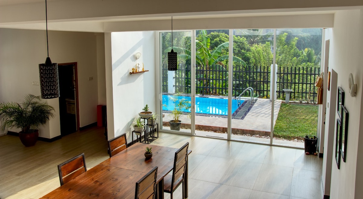 A spacious dining area features a wooden table surrounded by several chairs. Large glass panels provide a view of a backyard with a pool, greenery, and a wooden fence. Natural light fills the room, enhancing the open and airy atmosphere.