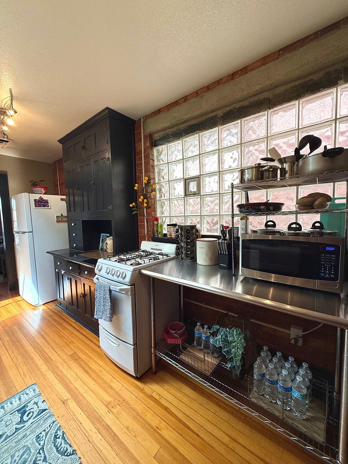 The kitchen space features a mix of modern and vintage elements, with a stainless steel countertop and a gas stove. A brick wall enhances the character, while sunlight filters through a glass block window. Shelving is filled with kitchen essentials and bottles of water.