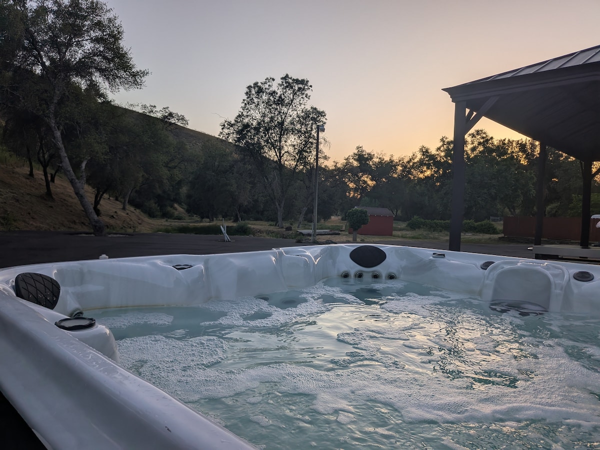 A spacious hot tub designed for eight guests sits under a canopy, with bubbling water visible. The surrounding landscape features trees and a gentle slope, bathed in warm hues from the setting sun.
