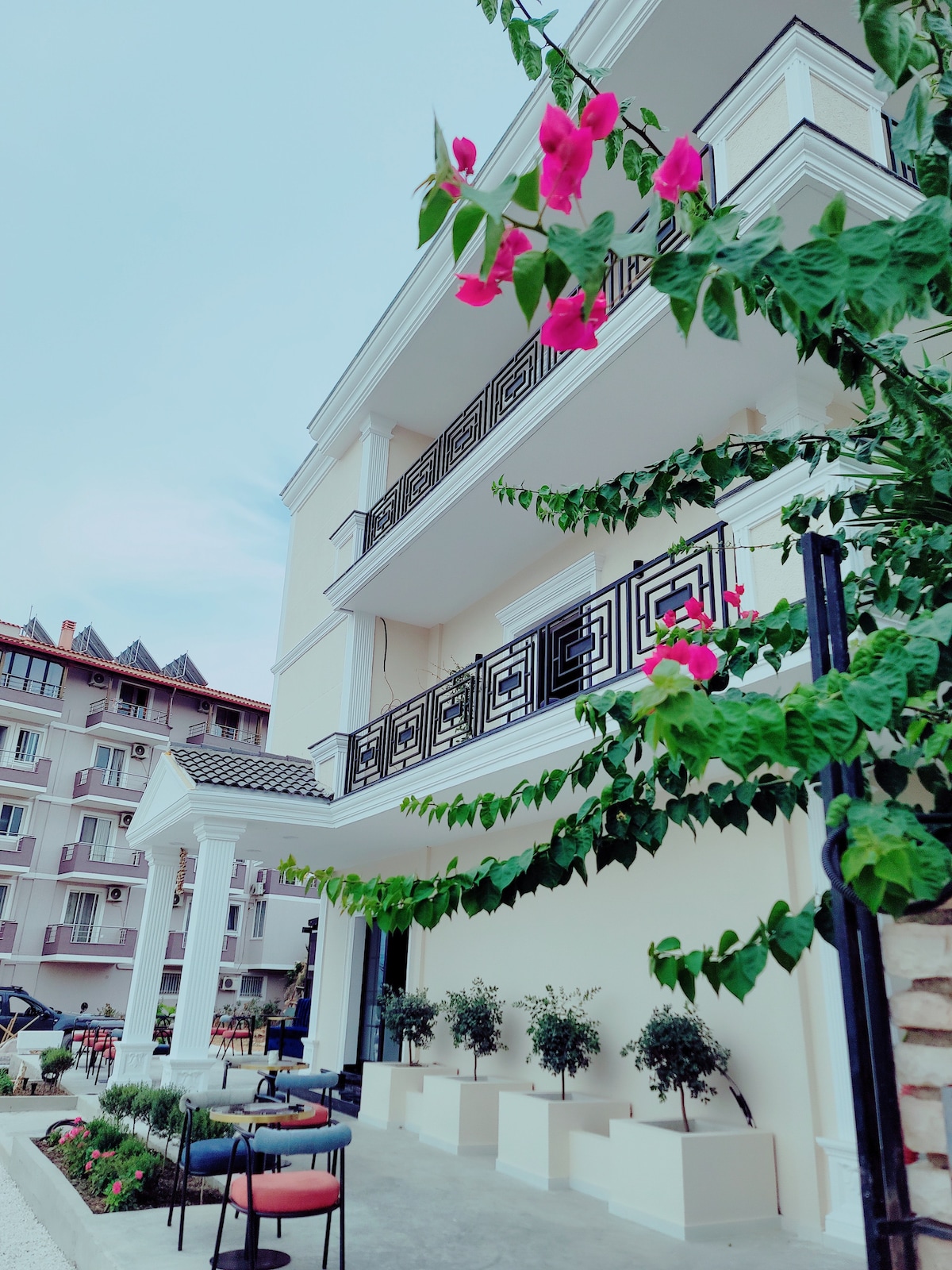 A stylish facade of the building is complemented by blooming flowers and greenery. Multiple balconies with decorative railings offer outdoor seating options, while planters with small trees line the area. The elegant structure is set against a soft blue sky.