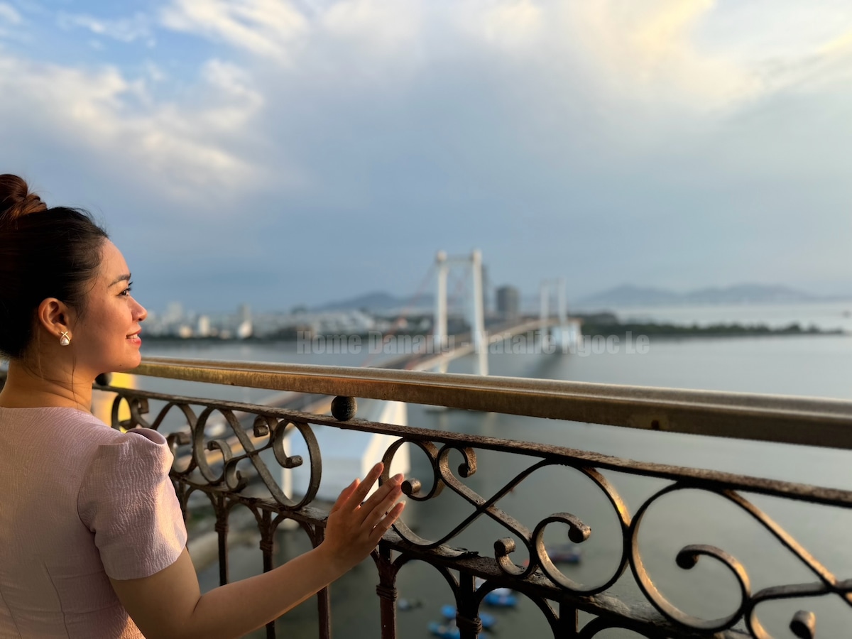 A captivating view from a high balcony showcases the ocean and a bridge in the distance. Intricate iron railing frames the scene, while the soft colors of the sky suggest a tranquil evening. The expansive landscape offers a sense of openness and serenity.