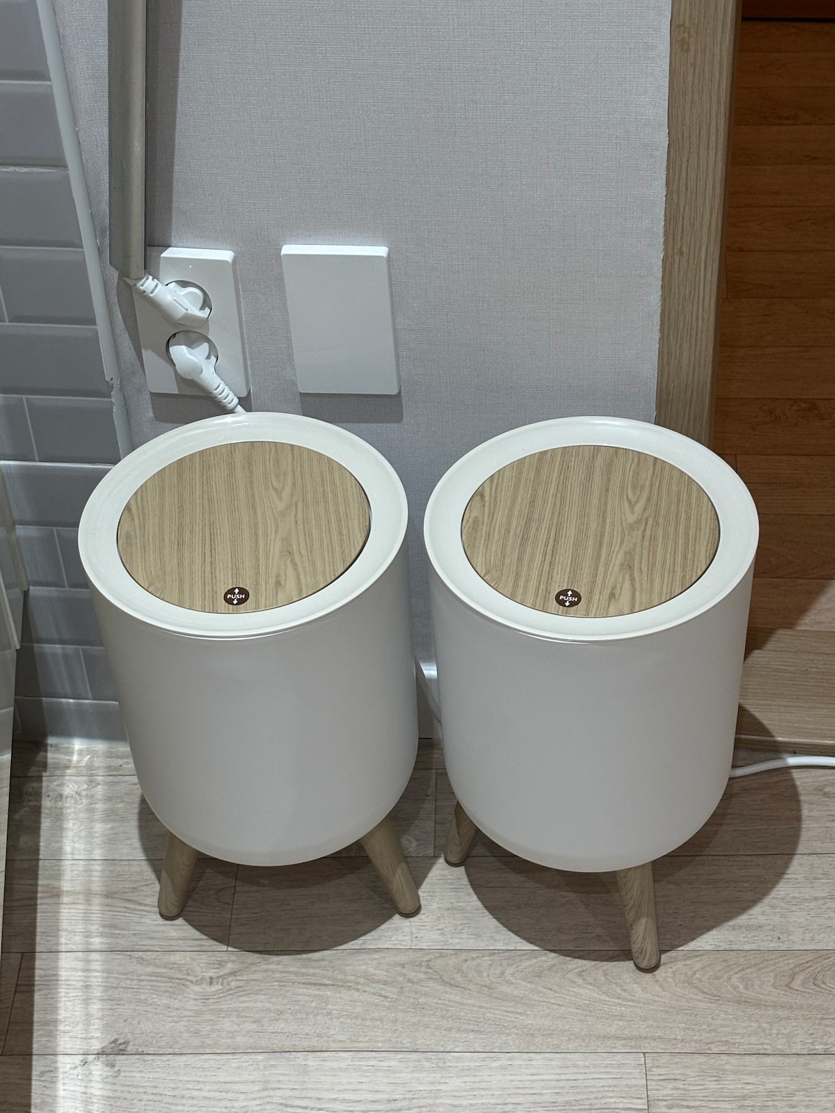 Two cylindrical trash bins are positioned side by side against a wall. Each bin features a white body with wooden lids, resting on short, wooden legs. The electrical outlets and cords are visible next to the bins, contributing to the functional design of the space.
