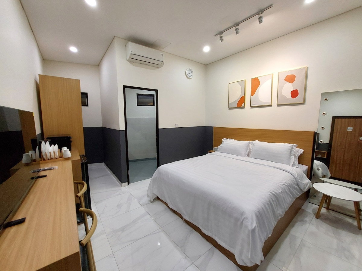 A modern room features a large queen bed with crisp white linens. A minimalist desk and seating area are situated along one wall. Natural light fills the space, highlighting the artwork above the bed and the sleek cabinetry. An air conditioning unit is installed for comfort.