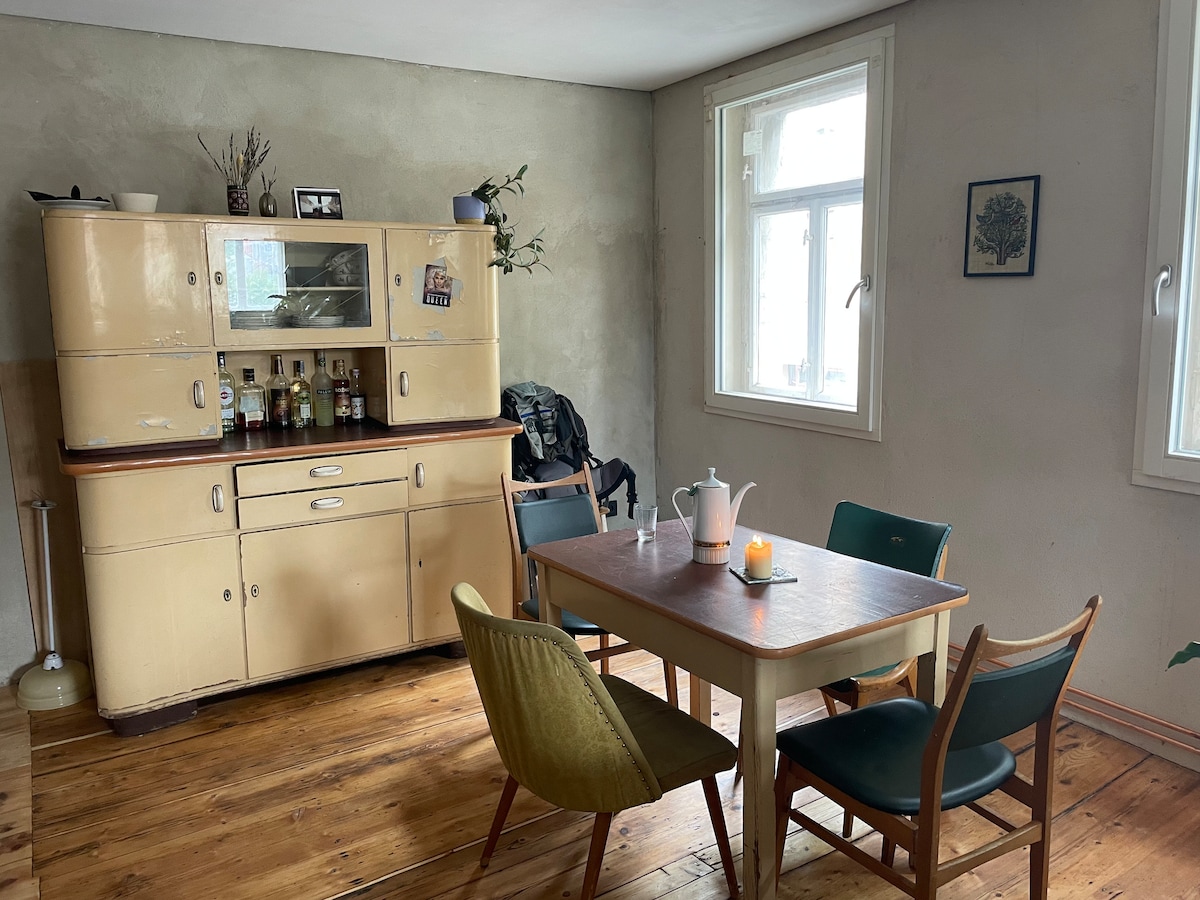 A light-filled kitchen features a vintage cabinet displaying a variety of glass bottles. A wooden table is surrounded by four chairs in differing colors, with a kettle and candle atop the table. Large windows provide natural light to the space.
