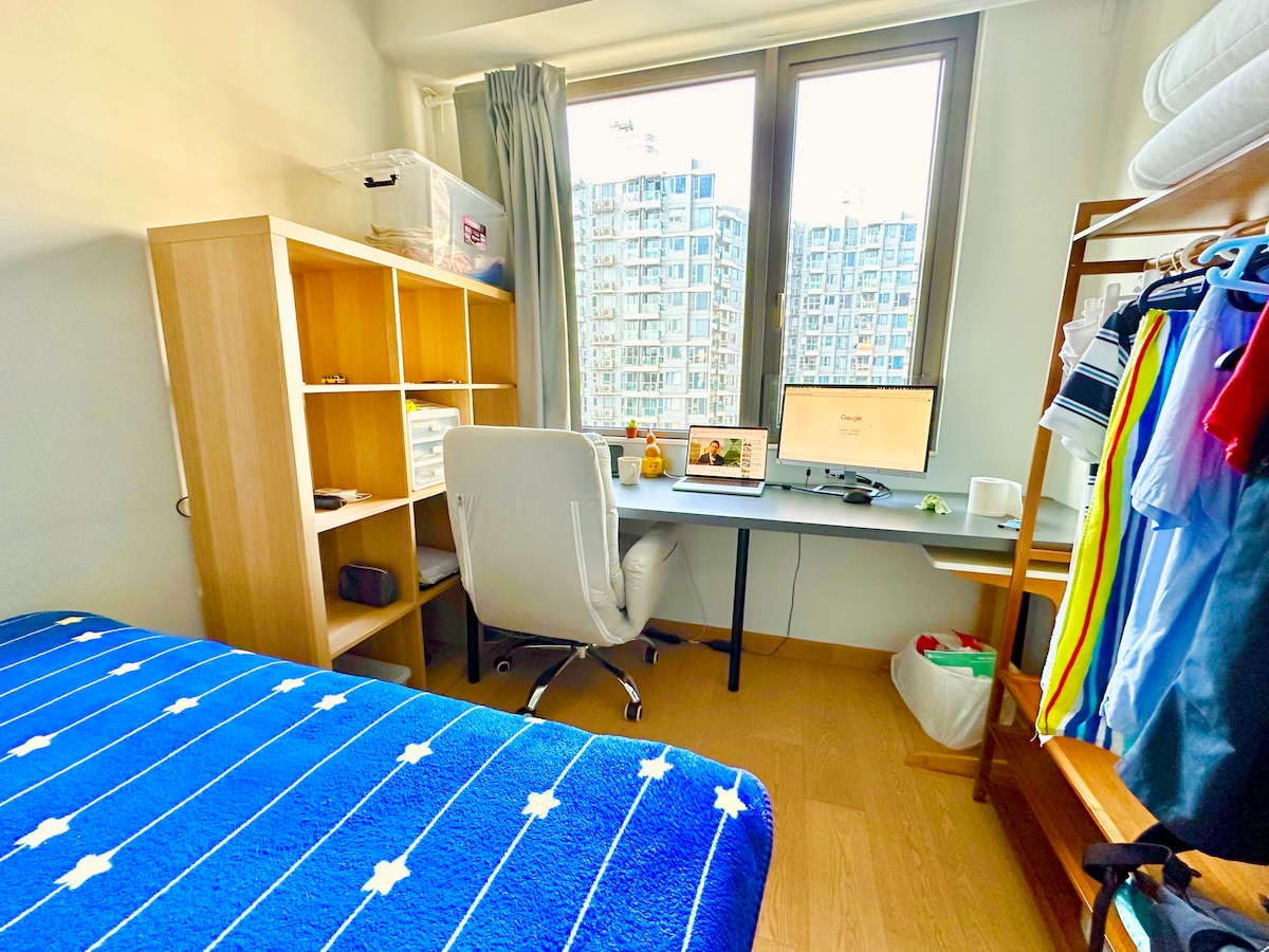 The spacious master bedroom features a queen-size bed with a blue star-patterned blanket. A desk with a computer and a white chair is positioned near a large window, offering views of the city. Shelving units and a clothing rack add functionality to the space.