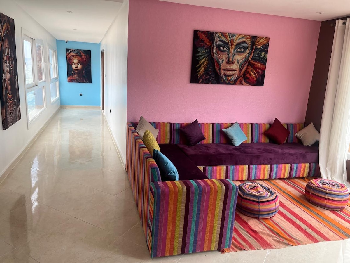 A vibrant lounge area is highlighted by a colorful striped sofa adorned with decorative pillows. The walls are painted in soft hues of pink and blue, creating a welcoming atmosphere. Natural light floods in through large windows, illuminating the space and its polished floor.