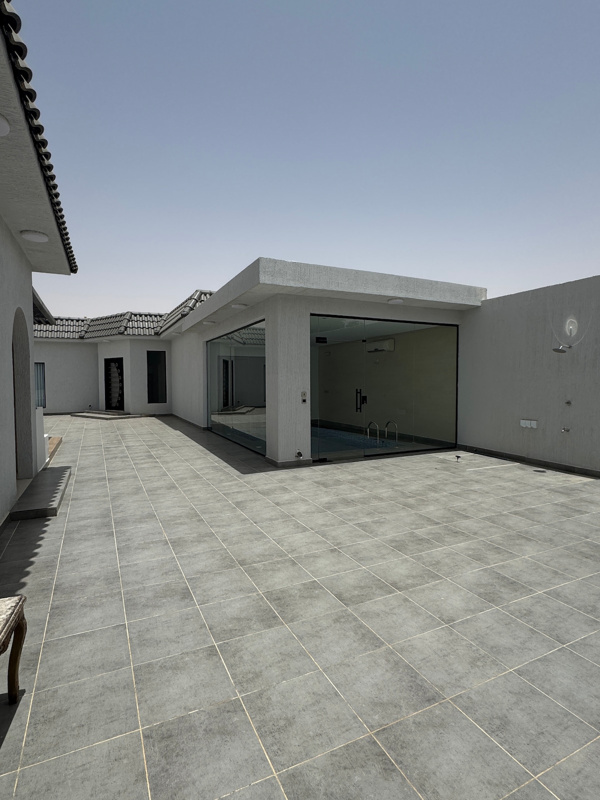 An outdoor terrace area is showcased, featuring large gray tiles that extend throughout the space. A glass wall reveals an indoor area, providing a view of the swimming pool. The surrounding architecture is characterized by clean lines and a neutral color palette.
