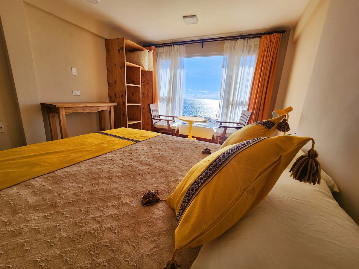 A spacious sleeping area is highlighted by a large bed adorned with vibrant yellow accents and decorative pillows. Natural light fills the room through large windows, which offer a view of the lake and nearby seating for two.