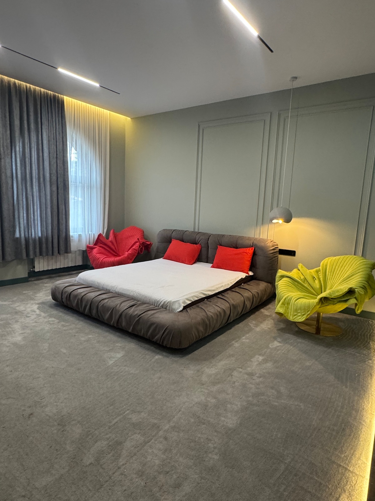 A spacious bedroom is highlighted by a low-set brown bed adorned with white linens and red accent pillows. Two modern chairs in vibrant green and pink add a pop of color. Soft gray curtains frame a window, allowing natural light to brighten the room.