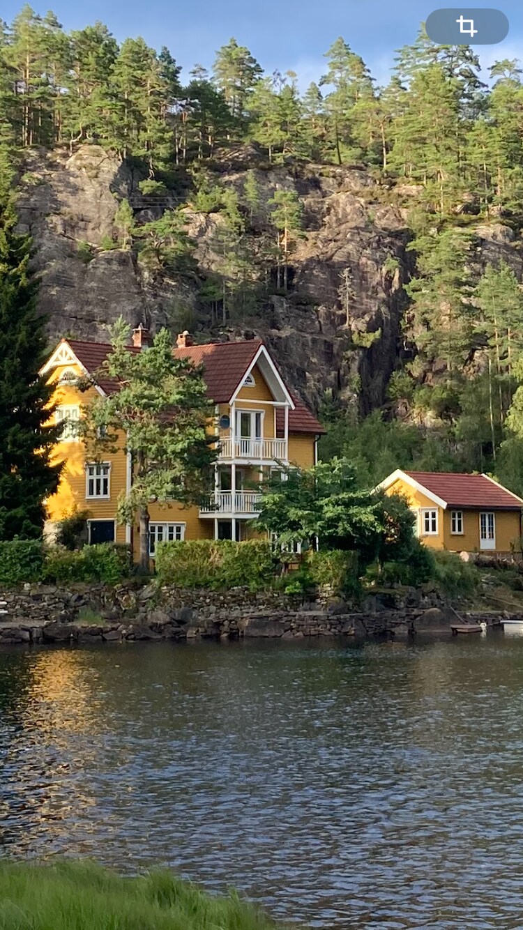 A charming yellow house is situated by a calm body of water, framed by lush greenery and towering cliffs in the background. Large windows provide views of the surrounding landscape. The peaceful scene suggests a serene retreat away from urban life.
