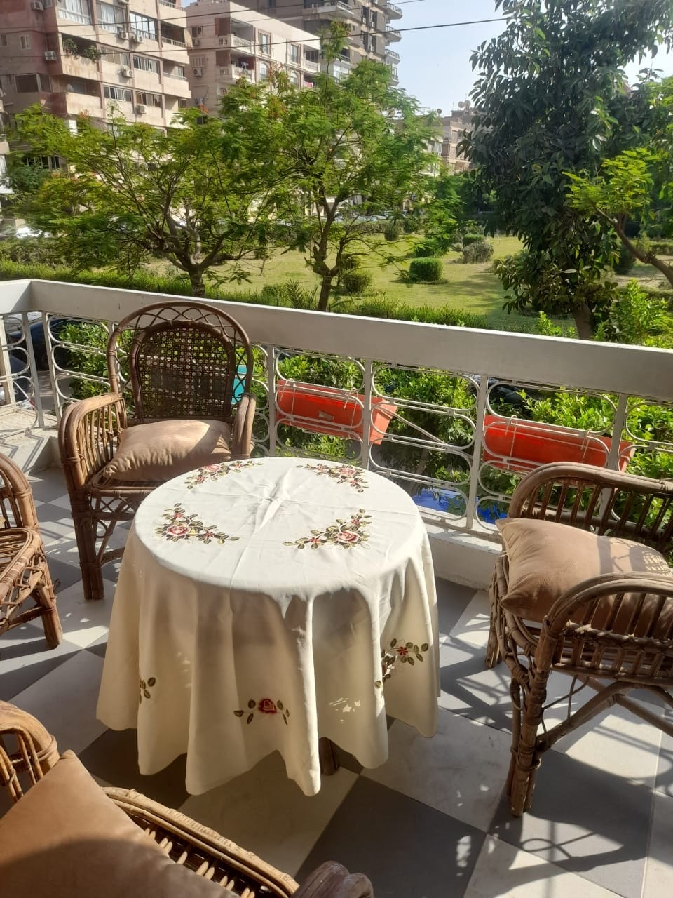 A charming balcony area features a round table draped with an embroidered tablecloth surrounded by four woven chairs. Lush greenery is visible in the background, complemented by colorful flower pots that add vibrant touches to the peaceful view.