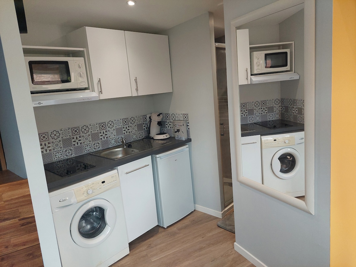 A compact kitchen area is displayed, featuring white cabinetry and modern appliances, including a microwave and a washing machine. The counter is adorned with decorative tiles, while a mirror reflects the space, enhancing its sense of openness.