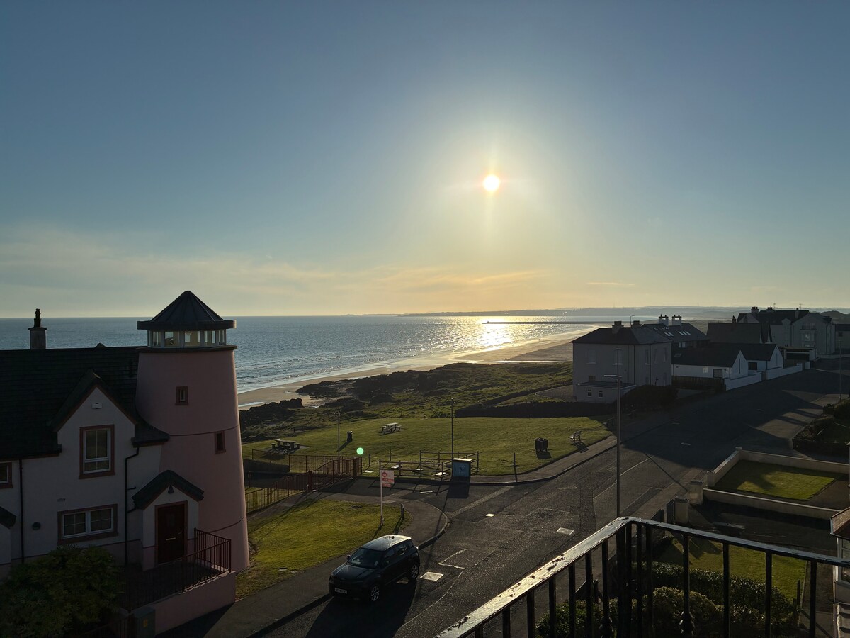 A sunlit landscape features a view of the Atlantic Ocean, with shimmering water reflecting natural light. The beach is visible in the foreground, framed by a grassy area and distant structures along the coastline.