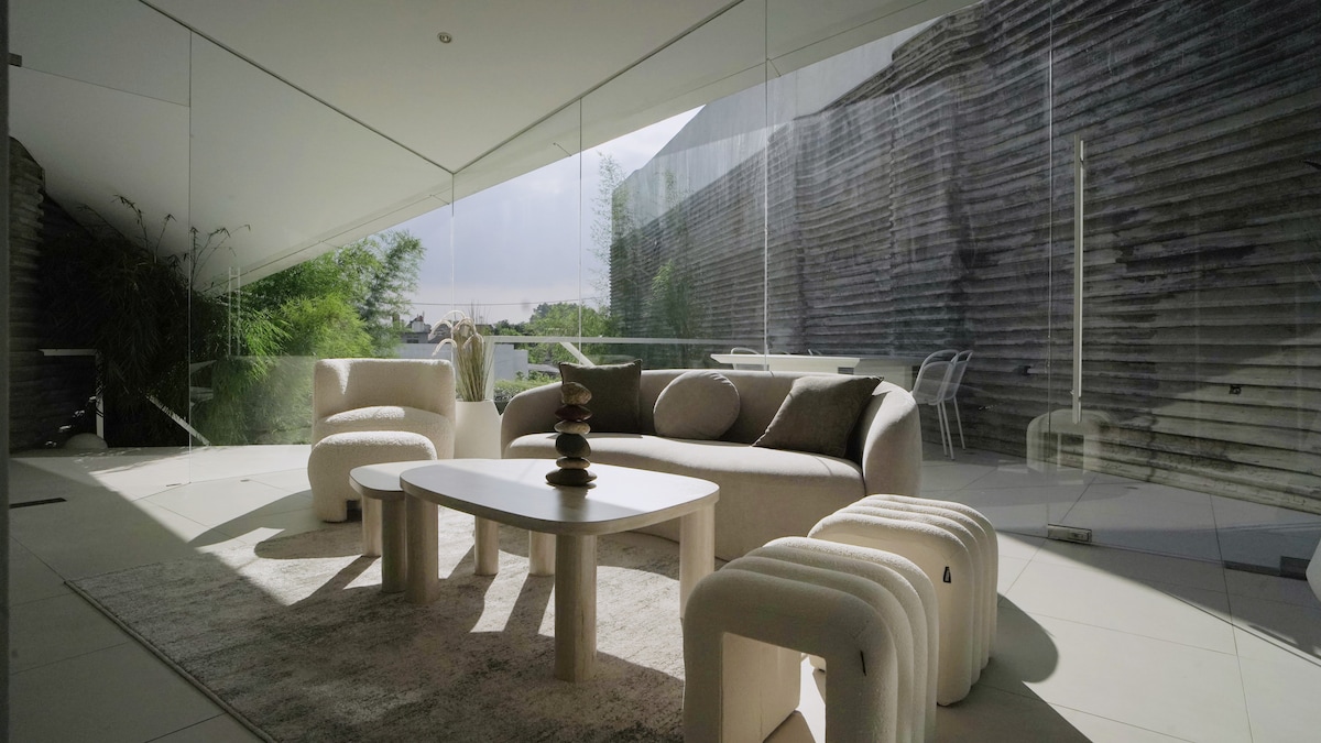 A contemporary living area is featured with a cozy sofa and armchairs, arranged around a minimalist coffee table. Natural light floods the space through large glass panels, highlighting the elegant decor and soft-colored furnishings. A serene outdoor area with greenery is visible through the glass.