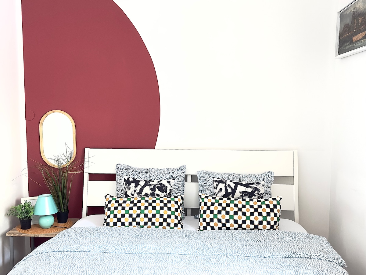A neatly arranged sleeping area features a spacious bed with patterned cushions and a light blanket. A round mirror and a small lamp are placed on a bedside table, accompanied by a decorative plant. A bold accent wall adds a pop of color.