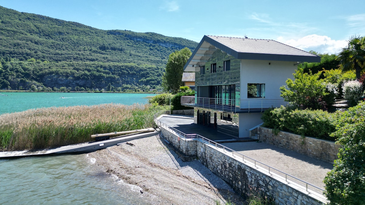 The exterior of the villa is shown from the lakeside, featuring a modern architectural design with large glass windows. A private beach area is visible in front, leading to the turquoise waters of the lake, framed by nearby lush vegetation and mountains in the background.