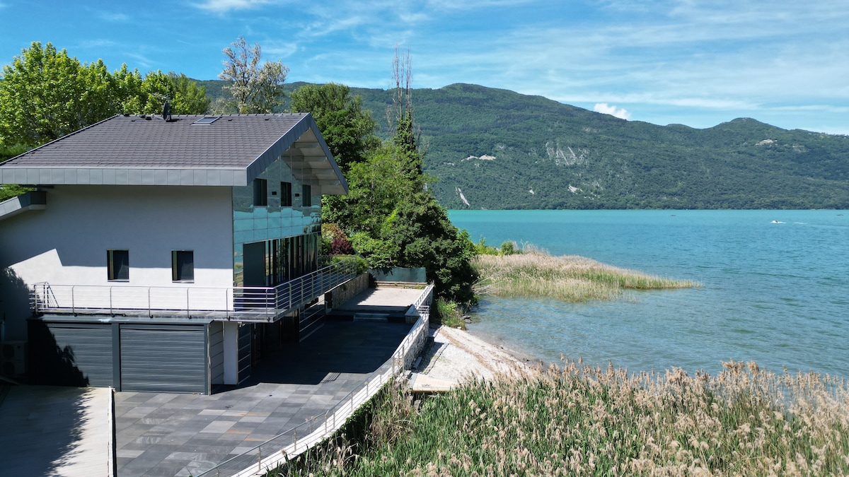A modern villa is set against the backdrop of a serene lake and lush mountains. Expansive glass walls reflect the surrounding nature, while a spacious outdoor terrace extends towards the water's edge, bordered by tall grasses and gentle waves.
