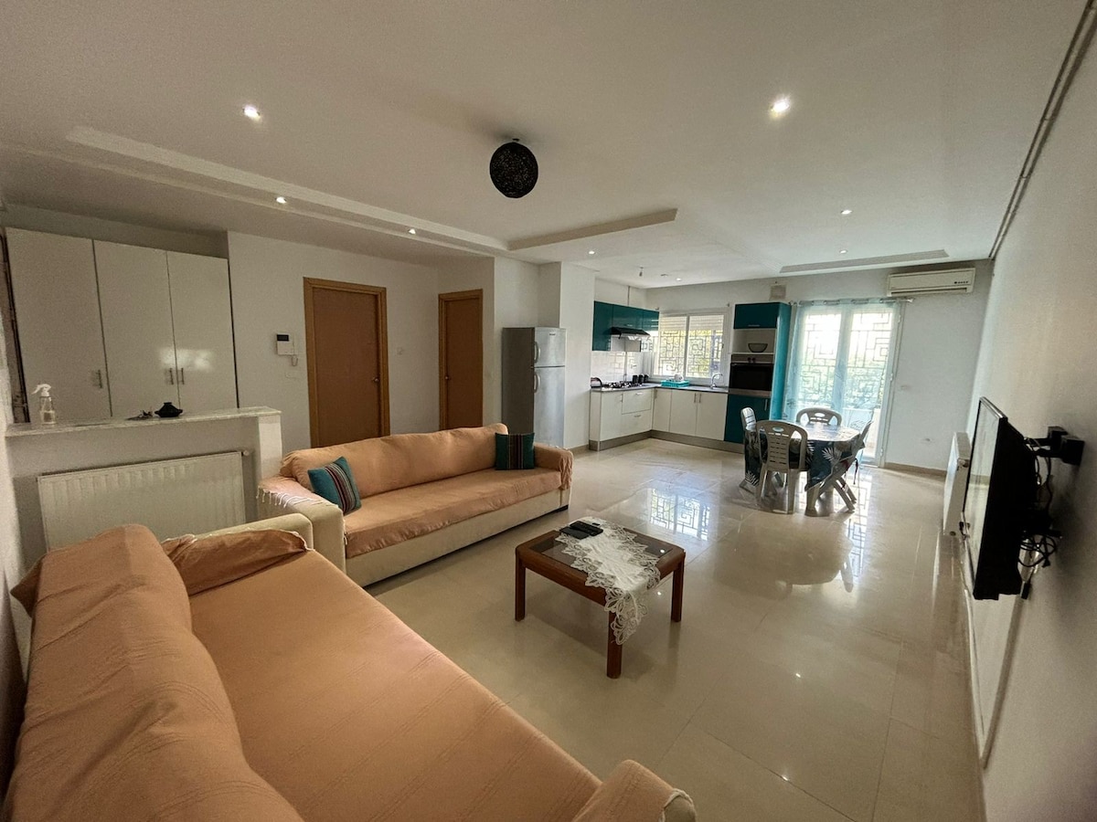 A spacious living area is showcased with two light-colored sofas positioned around a wooden coffee table. The kitchen is visible in the background, featuring modern appliances and a dining table surrounded by chairs. Natural light fills the room through large windows.