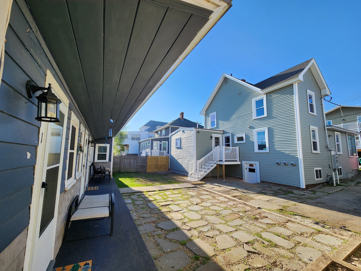 A spacious outdoor area is framed by two buildings, featuring a stone-paved pathway and a small patio. One building displays a covered porch with seating, while the other showcases a staircase leading to an upper level. Natural light enhances the inviting atmosphere.