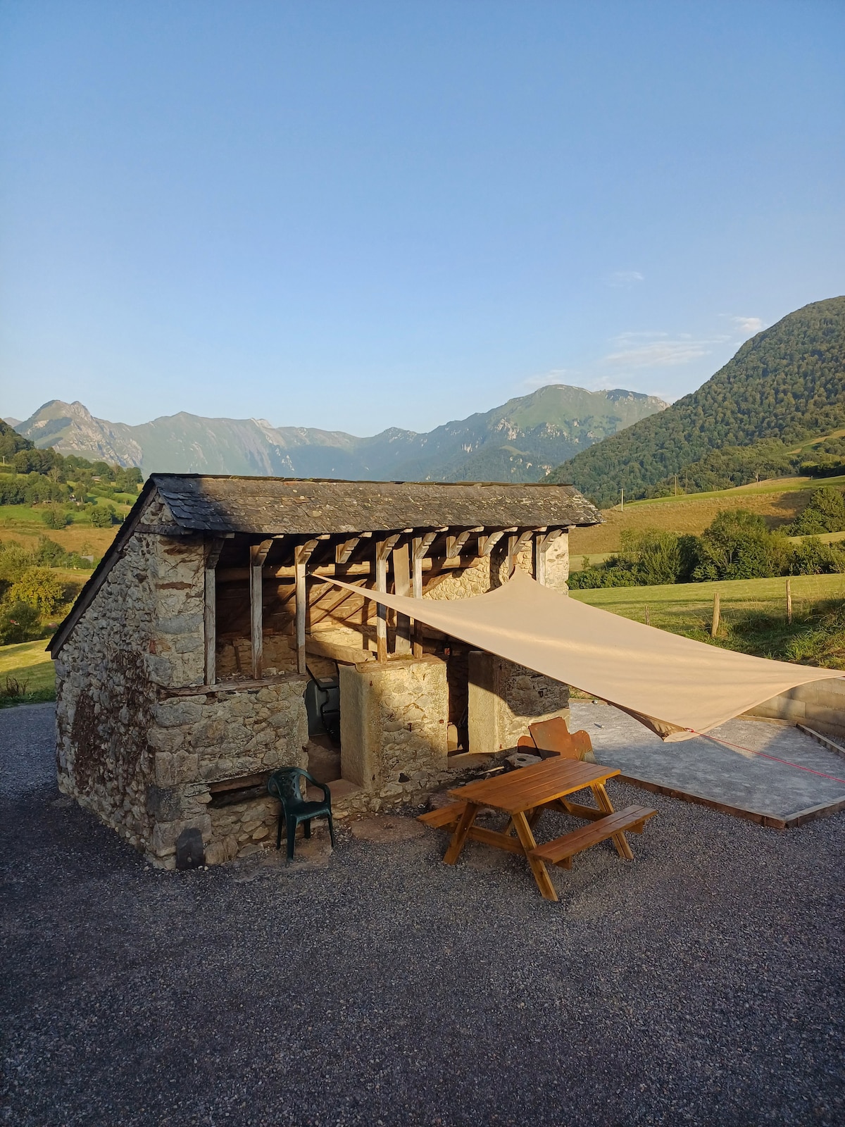 A charming outdoor space is featured, showcasing a rustic stone building with a sloping roof. A large canvas shade extends from the building, providing cover for a wooden picnic table and chairs. Scenic mountain views are visible in the background, enhancing the tranquil setting.