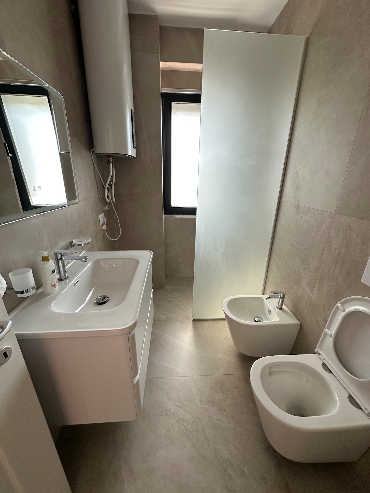 A modern bathroom is presented with neutral tiled walls and floors. A sleek vanity with a sink and large mirror is visible, alongside a shower area separated by a glass panel. A toilet and bidet are situated opposite, with natural light entering through a window.